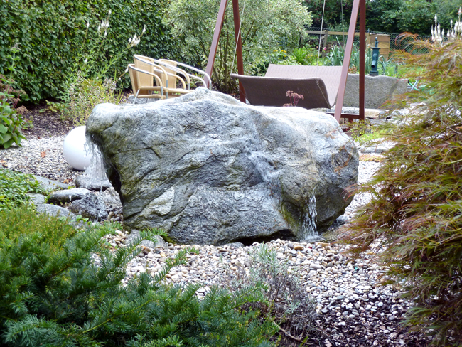 Quellstein mit Wasserlauf und im Hintergrund Schaukel im Kiesbeet